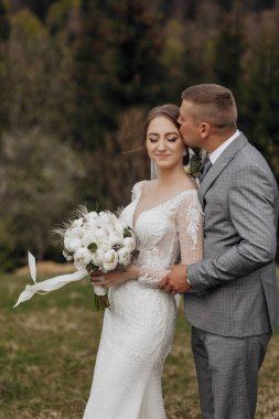 Bir gelin ve damat tarlada birbirlerine sarılıyorlar. Gelin beyaz bir elbise giyiyor ve elinde bir buket çiçek tutuyor. Sahne romantik ve samimi, aşkı yakalıyor.