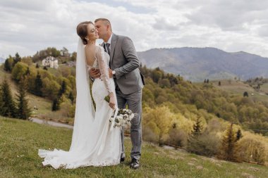 Bir gelin ve damat çimenli bir yamaçta öpüşüyor. Gelin beyaz bir elbise giyiyor ve damat bir takım elbise giyiyor. Sahne huzurlu ve romantik.