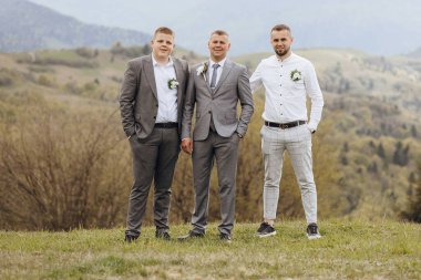 Takım elbiseli üç adam çimenli bir yamaçta durup fotoğraf için poz veriyor. Adamlar resmi giysiler giymişler, biri kravat takmış. Sahnenin rahat ve rahat bir havası var.