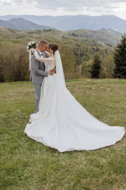 Gelin ve damat bir tarlada duruyorlar, gelin beyaz bir elbise giyiyor ve damat takım elbise giyiyor.