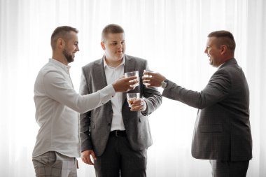 Üç adam bir odada duruyor, her biri bir bardak içki tutuyor. Hepsi gülümsüyor ve birbirlerinin arkadaşlığından zevk alıyor gibi görünüyorlar. Sahne neşeli ve sosyaldir, erkekler içkisini paylaşırken.