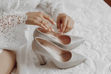 Bir kadın yüksek topuklu ayakkabıları ile bir yatakta oturuyor. O, beyaz bir elbise giyiyor ve elleri ayaklarının üzerinde. Zarafet ve incelik kavramı