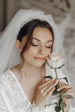 Bir kadın elinde bir gül tutuyor ve ona bakıyor. Beyaz bir gelinlik ve duvak giyiyor.