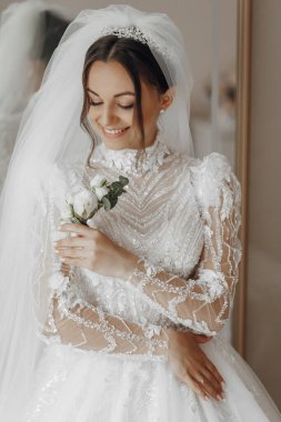 Bir gelin beyaz bir elbise giyiyor ve elinde bir çiçek tutuyor. Gülümsüyor ve mutlu.