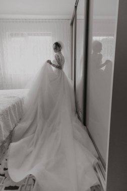 Gelinlikli bir kadın aynanın önünde duruyor. Elbise uzun ve dalgalı ve kadın bir peçe tutuyor. İmajın romantik ve zarif bir havası var.