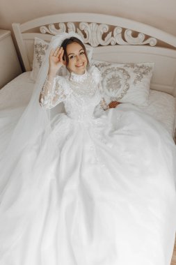 Beyaz elbiseli bir yatakta oturan bir gelin. Gülümsüyor ve elini kaldırıyor.
