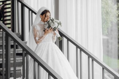 Bir gelin merdivende elinde bir buket çiçekle dikiliyor. Gelin beyaz bir elbise ve duvak takıyor. Merdiven metalden yapılmıştır ve beyaz perdeli bir odadadır.