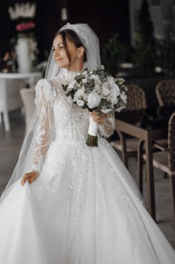 Bir gelin uzun beyaz bir elbise giyiyor ve elinde bir buket çiçek tutuyor. Sandalyeli bir masanın önünde duruyor.