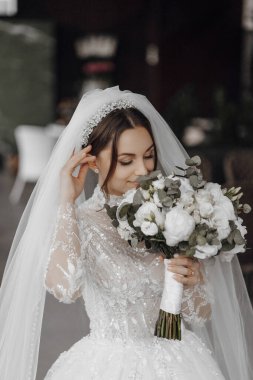 Bir gelin elinde bir buket beyaz çiçek ve duvak tutuyor. Beyaz bir elbise ve taç giyiyor.