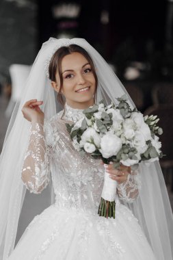 Bir gelin beyaz bir elbise giyiyor ve elinde bir buket çiçek tutuyor. Gülümsüyor ve kameraya bakıyor.