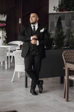 Takım elbiseli bir adam elinde bir vazo çiçekle bir masanın önünde duruyor. O bir kravat ve siyah bir takım elbise giyiyor.