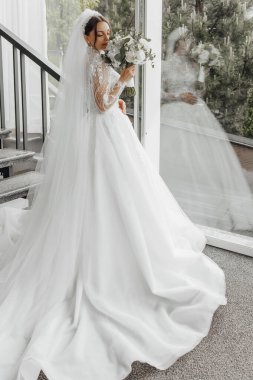 Bir gelin beyaz bir gelinlik giyiyor ve elinde bir buket çiçek tutuyor. Elbise uzun ve dalgalı ve gelin bir merdivenin üzerinde duruyor. Zarafet ve kutlama kavramı