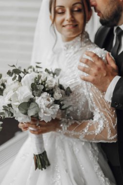 Gelin ve damat kucaklaşırken gelin bir buket çiçek tutuyor. Gelin beyaz bir elbise giyiyor ve damat bir takım elbise giyiyor. Sahne bir düğün fotoğrafı.