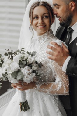 Gelin ve damat birbirlerine sarılıyorlar, gelin de bir buket çiçek tutuyor. Gelin beyaz bir elbise giyiyor ve damat bir takım elbise giyiyor. Sahne mutlu ve romantik.
