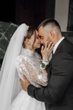 Bir gelin ve damat birbirlerinin yüzlerini öpüyor. Gelin beyaz bir elbise giyiyor ve damat da takım elbise giyiyor.