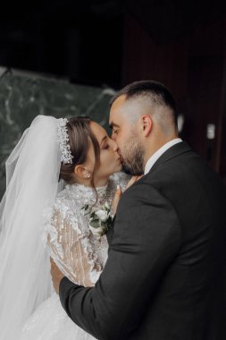 Yeşil bir duvarın önünde bir gelin ve damat öpüşüyor. Gelin beyaz bir elbise ve duvak takıyor.