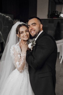 Bir gelin ve damat fotoğraf için poz veriyor. Gelin beyaz bir elbise giyiyor ve damat siyah bir takım elbise giyiyor. İkisi de gülümsüyor ve mutlu görünüyorlar.