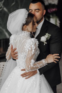 Bir gelin ve damat bir düğün töreninde birbirlerini kucaklıyorlar. Gelin beyaz bir elbise giyiyor ve damat siyah bir takım elbise giyiyor. Sahne aşk ve mutluluktur.