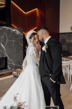 Gelin ve damat, gelin beyaz elbise ve damat siyah takım elbise giyerek bir odada yan yana duruyorlar. Birbirlerine gülümsüyor ve sarılıyorlar, mutlu ve mutlu görünüyorlar.