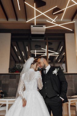 Bir gelin ve damat birbirlerine yakın duruyorlar, gelin uzun bir elbise giyiyor ve damat takım elbise giyiyor. Sahne romantik ve samimi, çünkü çift birbirini kucaklıyor.
