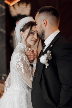 Bir gelin ve damat düğün fotoğrafında birbirlerine sarılıyorlar. Gelin beyaz bir elbise ve duvak giyerken damat takım elbise ve kravat takıyor. Fotoğraf bir aşk anını yakalar.