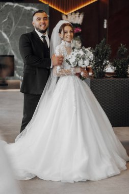 Bir gelin ve damat yeşil bir bitkinin önünde poz veriyorlar. Gelin beyaz bir elbise giyiyor ve damat bir takım elbise giyiyor. Gelin elinde bir buket çiçek tutuyor.