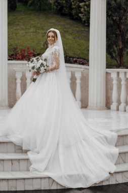 Beyaz gelinlikli bir kadın mermer bir merdivenin üzerinde duruyor. O, elinde bir buket çiçek tutuyor. Gelin güzel bir elbise giydiği için, zarafet ve kutlama kavramı.