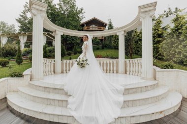 Bir gelin elinde bir buket çiçekle beyaz bir kemer yolunun önünde duruyor. Kemer yolu sütunlarla çevrili ve gelin uzun beyaz bir elbise giyiyor. Sahne zarif ve romantik.