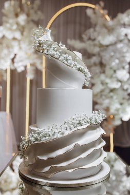 Arkasında beyaz kemer olan beyaz bir düğün pastası. Pasta uzun ve üç katlı.