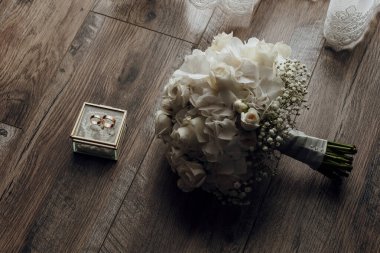 Tahta bir zeminde küçük bir kutunun yanında bir buket beyaz çiçek duruyor. Kutuda iki yüzük var. Düğün hediyesi ya da aşkın sembolü olabilir. Romantizm ve kutlama kavramı