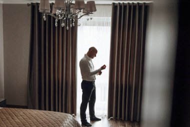 Bir adam pencerenin önünde duruyor, kravatını düzeltiyor. Oda loş bir şekilde aydınlanıyor ve perdeler çekiliyor, mahremiyet ve yakınlık hissi yaratıyor. Adam odaklanmış ve ciddi bir ruh hali içinde.