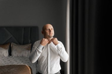 Beyaz gömlekli bir adam kravatını bağlamaya hazırlanıyor. O bir pencerenin önünde duruyor ve oda loş bir şekilde aydınlanıyor.