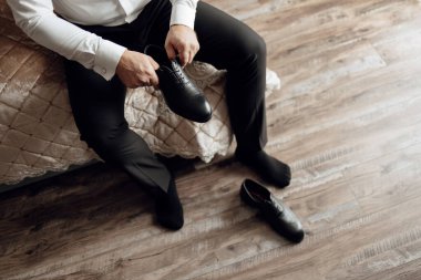 Bir adam yatakta oturuyor ve ayakkabılarını giyiyor. Ayakkabılar siyah ve adam siyah pantolon giyiyor.