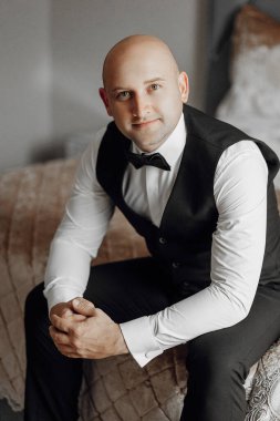 Siyah yelek ve siyah papyon takan bir adam yatağa oturur. Gülümsüyor ve kameraya bakıyor.