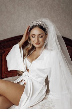 Beyaz bir elbise ve taç giymiş bir kadın bir yatakta oturuyor. Gülümsüyor ve mutlu.