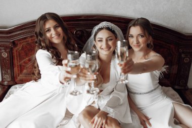 Üç kadın bir yatakta oturuyor, her biri bir bardak şarap tutuyor. Gülümsüyorlar ve özel bir olaya kadeh kaldırıyorlar. Sahne kutlama ve neşe dolu.