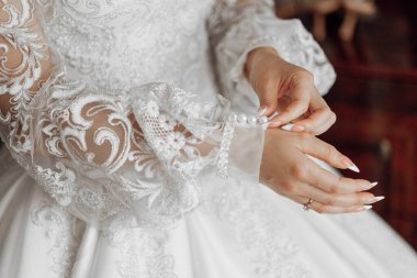 Bir kadın düğün için hazırlanıyor ve elbisesinin dantelini düzeltiyor. Elbise beyaz ve dantel bir yakası var. Kadın parmağına yüzük takmış.