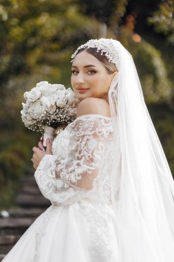 Bir gelin, bir buket beyaz çiçekle fotoğraf çektirmek için poz veriyor. Beyaz bir gelinlik ve beyaz bir duvak giyiyor. Mutluluk ve kutlama kavramı