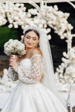 Bir gelin, bir buket beyaz çiçekle fotoğraf çektirmek için poz veriyor. Beyaz bir elbise ve duvak takıyor. Sahne arka planda beyaz kemerli bir bahçede geçiyor.