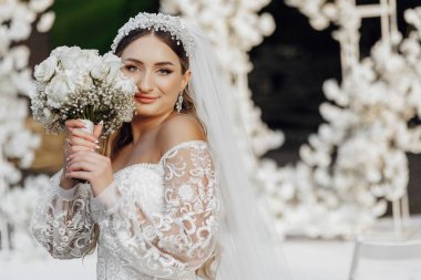 Bir gelin elinde bir buket beyaz çiçek tutuyor ve beyaz bir gelinlik giyiyor. Sahne arka planda beyaz kemerli beyaz bir odada geçiyor.