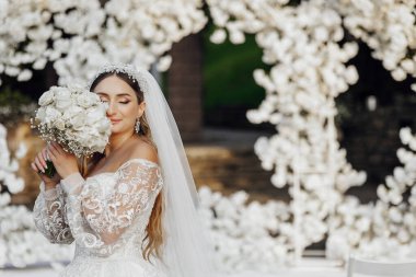 Bir gelin elinde bir buket beyaz çiçekle beyaz bir kemerin önünde duruyor.