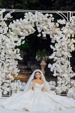 Beyaz bir kemerin önünde elinde çiçeklerle bir gelin oturuyor. Beyaz bir elbise ve duvak takıyor.