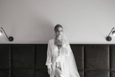 Beyaz elbiseli bir kadın siyah bir yatak başlığının önünde duruyor. Duvak ve kolye takıyor.