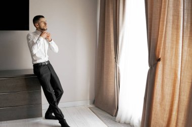 Takım elbiseli bir adam perdeli bir pencerenin önünde duruyor. Pencereden dışarı bakıyor, muhtemelen bir şeyler düşünüyor.