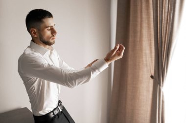 Beyaz gömlekli ve siyah pantolonlu bir adam perdenin önünde duruyor. Kolunu uzatıyor ve derin düşüncelere dalmış ya da rahatlamış bir ruh hali içinde.