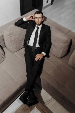 Takım elbiseli ve kravatlı bir adam kanepede oturuyor. O siyah bir takım ve beyaz bir gömlek giyiyor.