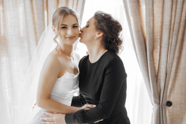 Bir kadın gelinin yanağından öper. Gelin beyaz bir elbise giyiyor.