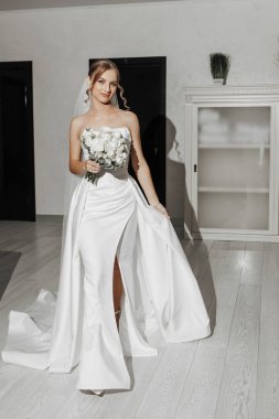 Bir gelin beyaz bir elbise giyiyor ve elinde bir buket çiçek tutuyor. Sahne zarif ve romantik.