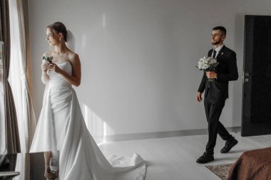 Bir gelin ve damat pencereli bir odada duruyorlar. Gelin bir buket tutuyor ve damat da bir buket tutuyor.
