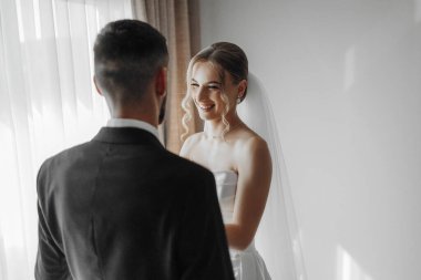 Bir gelin ve damat pencerenin önünde birbirlerine gülümsüyorlar. Gelin beyaz bir elbise giyiyor ve damat bir takım elbise giyiyor. Sahne mutlu ve neşeli.